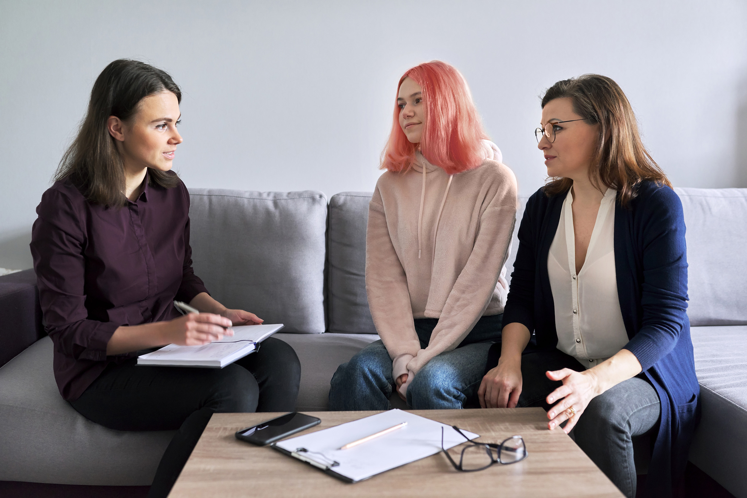 A counselor sits on a couch next to a young woman and her mother during a session